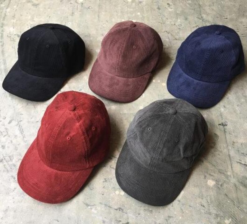 May include: Five baseball caps made of corduroy fabric, arranged in a group. The caps are colored in black, maroon, navy blue, bright red, and dark gray. Each cap features a rounded crown, a curved brim, and stitched eyelets on the panels.