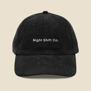 May include: A black corduroy baseball cap featuring the text Night Shift Co. embroidered in white thread on the front center. The cap has a structured crown, a curved brim, and a button at the top, showcasing a classic six-panel design.