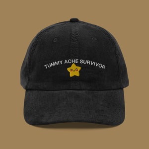 May include: Black corduroy baseball cap with the embroidered text "TUMMY ACHE SURVIVOR" above a yellow star with a smiling face. The cap has a curved brim and an adjustable strap.