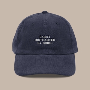 May include: A navy blue corduroy baseball cap with white embroidery that reads "EASILY DISTRACTED BY BIRDS".