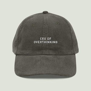 Può includere: Cappellino da baseball in velluto a coste verde oliva con la scritta ricamata "CEO OF OVERTHINKING" in bianco. Il cappellino ha una visiera curva e un cinturino regolabile. Il testo è centrato sul davanti.