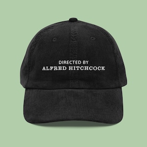 Op de afbeelding: Zwarte corduroy baseballpet met witte geborduurde tekst die "Directed by Alfred Hitchcock" luidt.