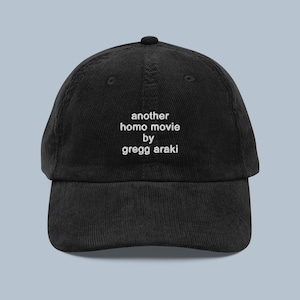 May include: Black corduroy baseball cap featuring the text "another homo movie by gregg araki" in white. The cap has a curved peak and an adjustable strap. A classic six-panel design.