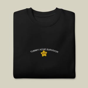 May include: A black crewneck sweatshirt featuring an embroidered design on the chest. The design includes the white text "TUMMY ACHE SURVIVOR" arched over a small, yellow, smiling star character with pink cheeks and closed eyes.