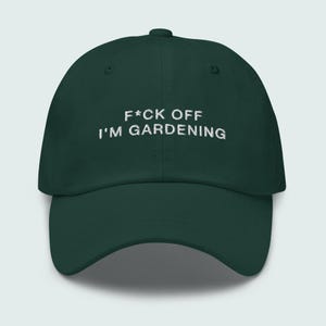 Könnte beinhalten: Dunkelgrüne Baseballkappe mit weiß gesticktem Text "F*ck Off I'm Gardening". Die Kappe hat einen gebogenen Schirm und einen Knopf oben. Ein humorvolles Accessoire für Gärtner.