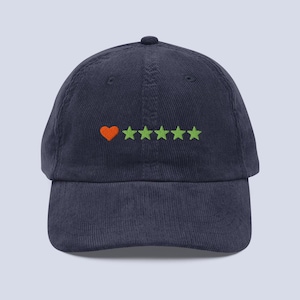 Puede incluir: Una gorra de béisbol de pana azul marino con un diseño bordado en la parte delantera. El bordado consiste en un corazón naranja seguido de cinco estrellas de color verde claro dispuestas en una fila horizontal en el centro.