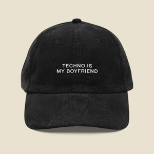 May include: Black corduroy baseball cap with the white text "TECHNO IS MY BOYFRIEND" embroidered on the front. The cap has a curved peak and a button on top. A casual accessory.