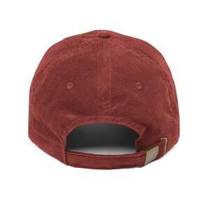 May include: A deep red corduroy baseball cap featuring a six-panel construction with stitched eyelets on the crown. The back of the cap includes an adjustable fabric strap with a metallic brass-toned buckle for a secure and customizable fit.