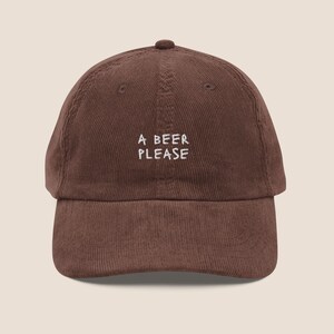 May include: A brown corduroy baseball cap featuring the embroidered white text A BEER PLEASE centred on the front panel. The hat has a structured crown, a curved brim, and stitched eyelets on the top panels for ventilation and a button top.