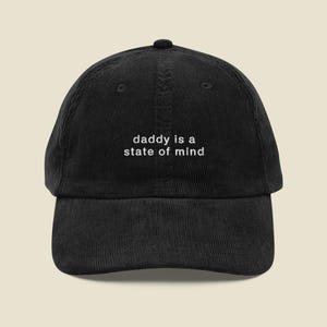 Könnte beinhalten: Schwarze Cord-Baseballkappe mit dem weißen Schriftzug "daddy is a state of mind" auf der Vorderseite. Die Kappe hat einen gebogenen Schirm und einen Knopf oben.
