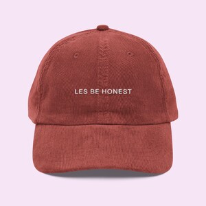 May include: A reddish-brown corduroy baseball cap with a curved peak and a rounded crown. The cap has the white embroidered text "LES BE HONEST" across the front. This casual cap is a stylish accessory for everyday wear.