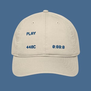 Pode incluir: Um boné de beisebol bege com texto bordado em azul que diz "PLAY 448C 0:88:0"