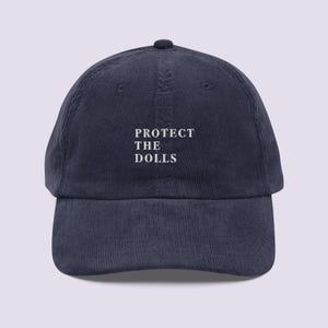 May include: A navy blue corduroy baseball cap with white embroidered text that reads "PROTECT THE DOLLS".