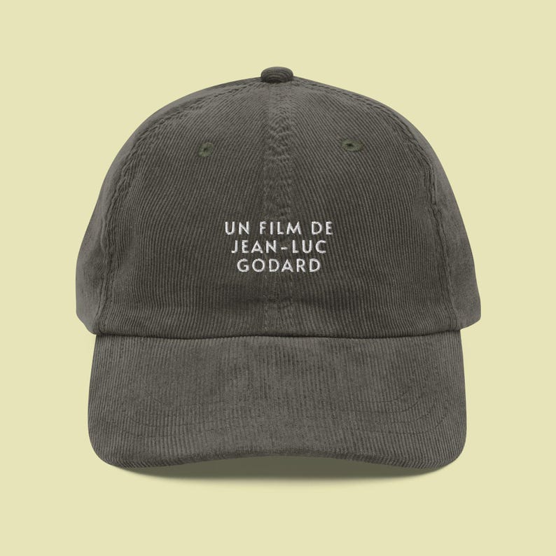 May include: A dark gray corduroy baseball cap with white embroidered text that reads "UN FILM DE JEAN-LUC GODARD".