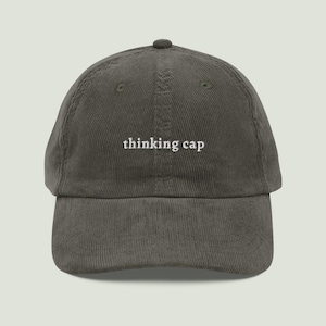 Pode incluir: Um boné de beisebol de veludo cotelê cinza-escuro com o texto "thinking cap" bordado em fio branco no painel frontal. O boné possui uma copa estruturada, aba curva e um botão no topo, apresentando um acabamento de tecido texturizado.