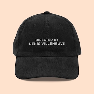 May include: Black corduroy baseball cap with white embroidered text that reads "DIRECTED BY DENIS VILLENEUVE".