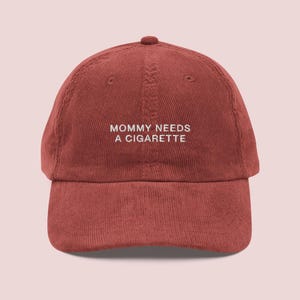 May include: A rust-colored corduroy baseball cap featuring the embroidered white text "MOMMY NEEDS A CIGARETTE" centered on the front panel. The hat has a structured crown, a curved brim, and visible vertical corduroy texture throughout the fabric.