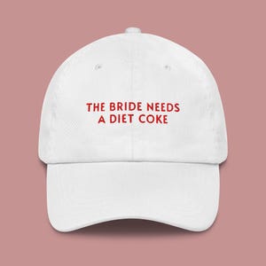 May include: White baseball cap with the red text "THE BRIDE NEEDS A DIET COKE". The cap has a curved brim and a fabric strap closure on the back. A fun accessory for a bachelorette party or wedding.
