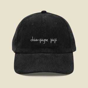 Può includere: Un cappellino da baseball in velluto a coste nero con il testo "champagne papi" ricamato in filo bianco sul pannello frontale. Il cappello presenta una corona strutturata, una visiera curva e un bottone al centro della parte superiore a sei pannelli.