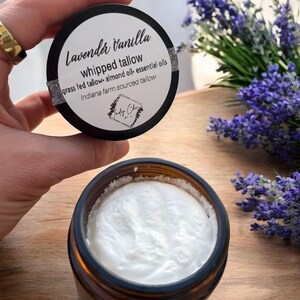 May include: A jar of Lavender Vanilla whipped tallow with a white, creamy texture. The jar has a black lid with white text and a logo. The ingredients include grass-fed tallow, almond oil, and essential oils. A sprig of purple flowers is in the background.