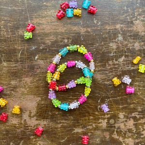 May include: Two circular bracelets made of translucent, multi-colored gummy bear-shaped beads and small metallic gold-tone spacer beads. Several loose gummy bear beads in shades of red, pink, yellow, green, blue, and purple are scattered around.