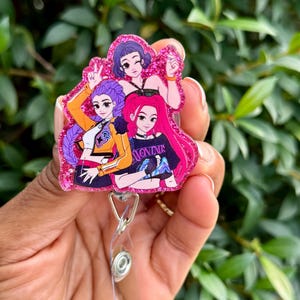 May include: A pink glitter badge reel with a cartoon illustration of three women with purple, pink, and black hair. The women are wearing different outfits. The badge reel has a clear plastic strap and a metal clip.