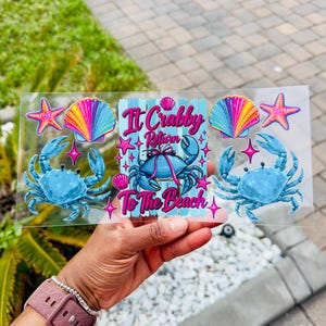 May include: A clear rectangular decal featuring two blue crabs, two pink starfish, and two rainbow-colored seashells. The center displays a blue wooden-plank background with a crab wearing a pink bow and the text It Crabby Return To The Beach.
