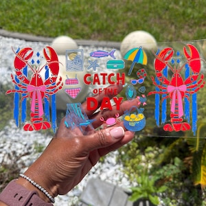 May include: A clear rectangular glass with a colorful beach-themed design. The design includes two red lobsters with pink bows, the text "CATCH OF THE DAY", and various beach icons like an umbrella, sunglasses, and a crab.