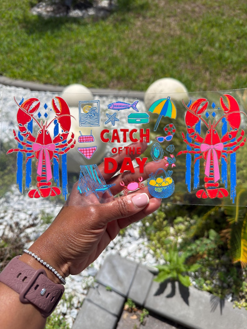 May include: A clear rectangular glass with a colorful beach-themed design. The design includes two red lobsters with pink bows, the text "CATCH OF THE DAY", and various beach icons like an umbrella, sunglasses, and a crab.