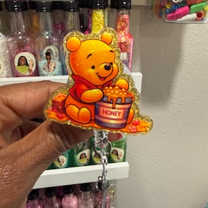 May include: A glittery acrylic badge reel featuring a yellow bear wearing a red shirt, sitting and eating from a blue pot labeled HONEY. The design includes small red heart accents and a golden sparkle finish, attached to a retractable cord mechanism.