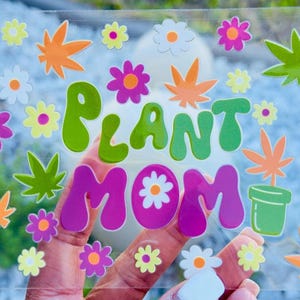 May include: A clear sticker with the words "PLANT MOM" in green and purple bubble letters, surrounded by colorful cannabis leaves, flowers, and a small pot. The design is vibrant and playful, perfect for plant lovers.