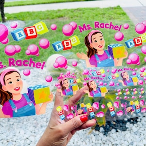 May include: A clear sticker sheet featuring multiple illustrations of a woman with brown hair wearing a pink headband, purple shirt, and blue overalls. The designs include the text Ms. Rachel, colorful ABC blocks, and various pink bubbles.