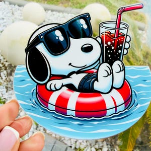May include: A Snoopy sticker featuring Snoopy wearing sunglasses and relaxing in a red and white striped inner tube in a pool. He is holding a glass of soda with a red straw. The sticker has a blue water background.