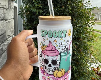 May include: A white frosted glass mug with a bamboo lid and a clear straw. The mug features a Halloween-themed design with a skull, pumpkins, and the words "Spooky & Sweet". The mug is held in a person's hand.