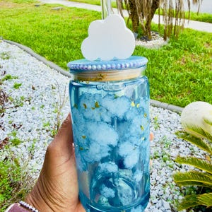 May include: A clear glass tumbler with a blue cloud design and a white cloud-shaped lid. The tumbler has a gold rim and a clear straw. The design includes gold flecks. The tumbler is held in a hand.