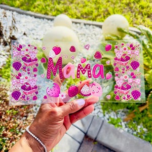 May include: A clear acrylic sign with the word "Mama" in pink, decorated with strawberries, bows, and hearts. The sign is held up by a hand, with a bracelet on the wrist. The background is blurred with greenery.