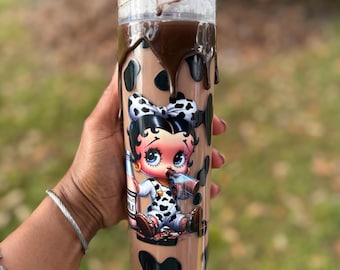 Könnte beinhalten: Ein klarer Becher mit einem schokoladenbraunen Design und schwarzen Kuhflecken. Der Becher zeigt ein Cartoon-Bild von Betty Boop. Die Oberseite ist mit Schlagsahne, Schokoladenguss und Streuseln gefüllt.
