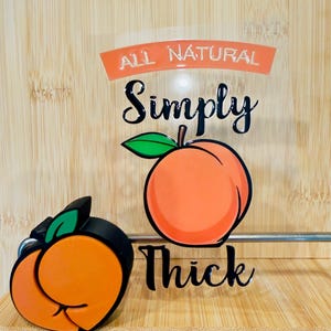 May include: A wooden shelf displays a peach-shaped keychain and a graphic with the words "ALL NATURAL Simply Thick" and a peach illustration. The keychain is orange, black, and green.