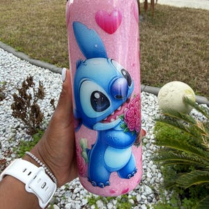 May include: A pink glitter tumbler with a blue cartoon character holding pink roses. The tumbler has a pink lid and a pink heart design. The character has large black eyes and a wide smile. The tumbler is designed for cold beverages.