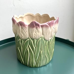 Może przedstawiać: Ceramiczna doniczka z motywem tulipanów. Doniczka jest biała z różowymi akcentami i zielonymi liśćmi.