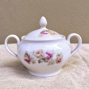 Op de afbeelding: Witte porseleinen suikerpot met deksel, versierd met een bloemenmotief van roze en gele rozen. De pot heeft een rond lichaam en twee handvatten.
