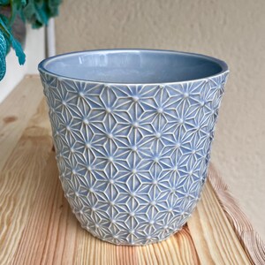 May include: A light blue ceramic planter with a geometric starburst pattern. The planter has a cylindrical shape and sits on a wooden surface. The interior of the planter is a solid light blue color.