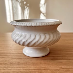 May include: White ceramic planter with a fluted design and a pedestal base. The planter is on a wooden surface.