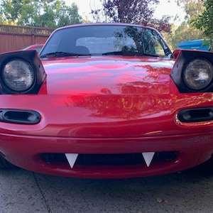 May include: A red Mazda Miata with black headlights and a white fang-like bumper trim. The car is parked on a gray asphalt surface.