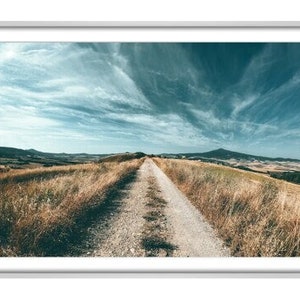 May include: A dirt road winds through a grassy field towards a distant mountain range under a blue sky with white clouds.