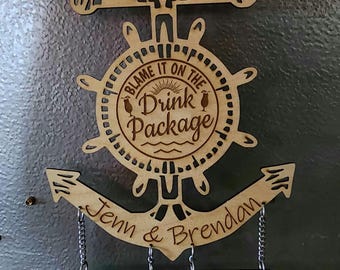 May include: A wooden anchor-shaped sign with the text "Blame it on the Drink Package" and the names "Jenn & Brendan" engraved on it.  The sign has three small wooden tags hanging from it with the text "Carnival Panorama April 8th, 2023 Mexican Riviera", "Carnival Miracle August 31st, 2023 Baja Mexico", and "Carnival Miracle April 6th, 2024 Hawaii 15 day cruise."
