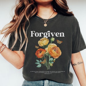 May include: A dark grey t-shirt with the word "Forgiven" printed in white. The shirt features a floral design with yellow and orange flowers and butterflies. The text "In Him we have redemption through His blood, the forgiveness of sins in accordance with the riches of God's grace Ephesians 1:7" is printed at the bottom of the shirt.