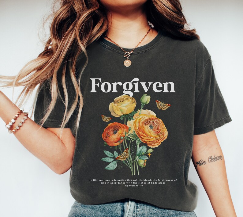 May include: A dark grey t-shirt with the word "Forgiven" printed in white. The shirt features a floral design with yellow and orange flowers and butterflies. The text "In Him we have redemption through His blood, the forgiveness of sins in accordance with the riches of God's grace Ephesians 1:7" is printed at the bottom of the shirt.