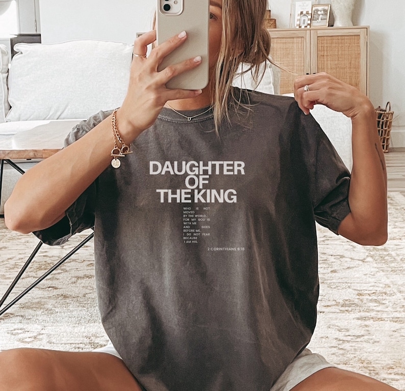 May include: A grey t-shirt with the text "Daughter of the King" and a Bible verse, 2 Corinthians 6:18, printed on the front.