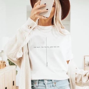 May include: A white short-sleeved t-shirt featuring a thin vertical black line forming a cross shape with the text "THE WAY, THE TRUTH, AND THE LIFE" printed horizontally across the center. The shirt is worn with a beige textured cardigan and blue jeans.